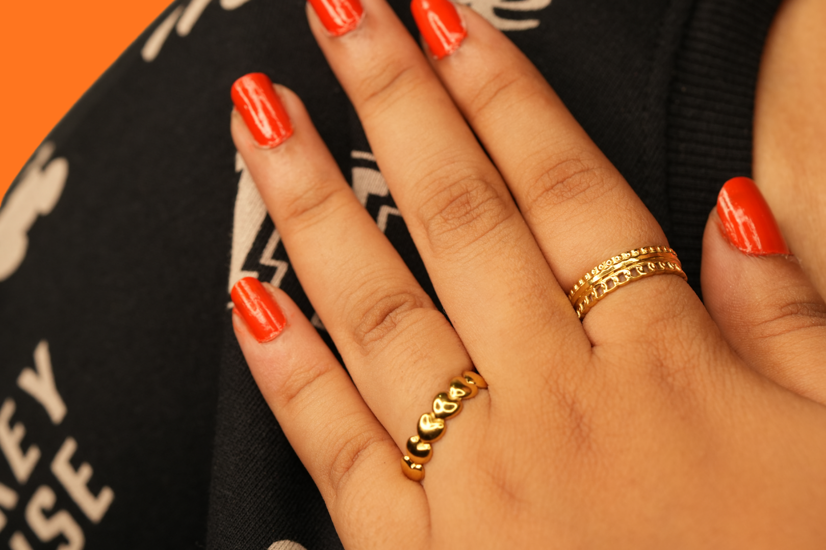 Close-up of a hand with red nail polish and 18k gold plated rings, wearing a black garment with white text.